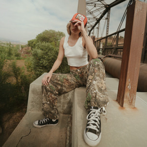 A casually dressed young person wearing camouflage pants, a white tank top, and a red cap sits on a concrete ledge of an urban bridge structure, adjusting their hat and looking slightly downward.